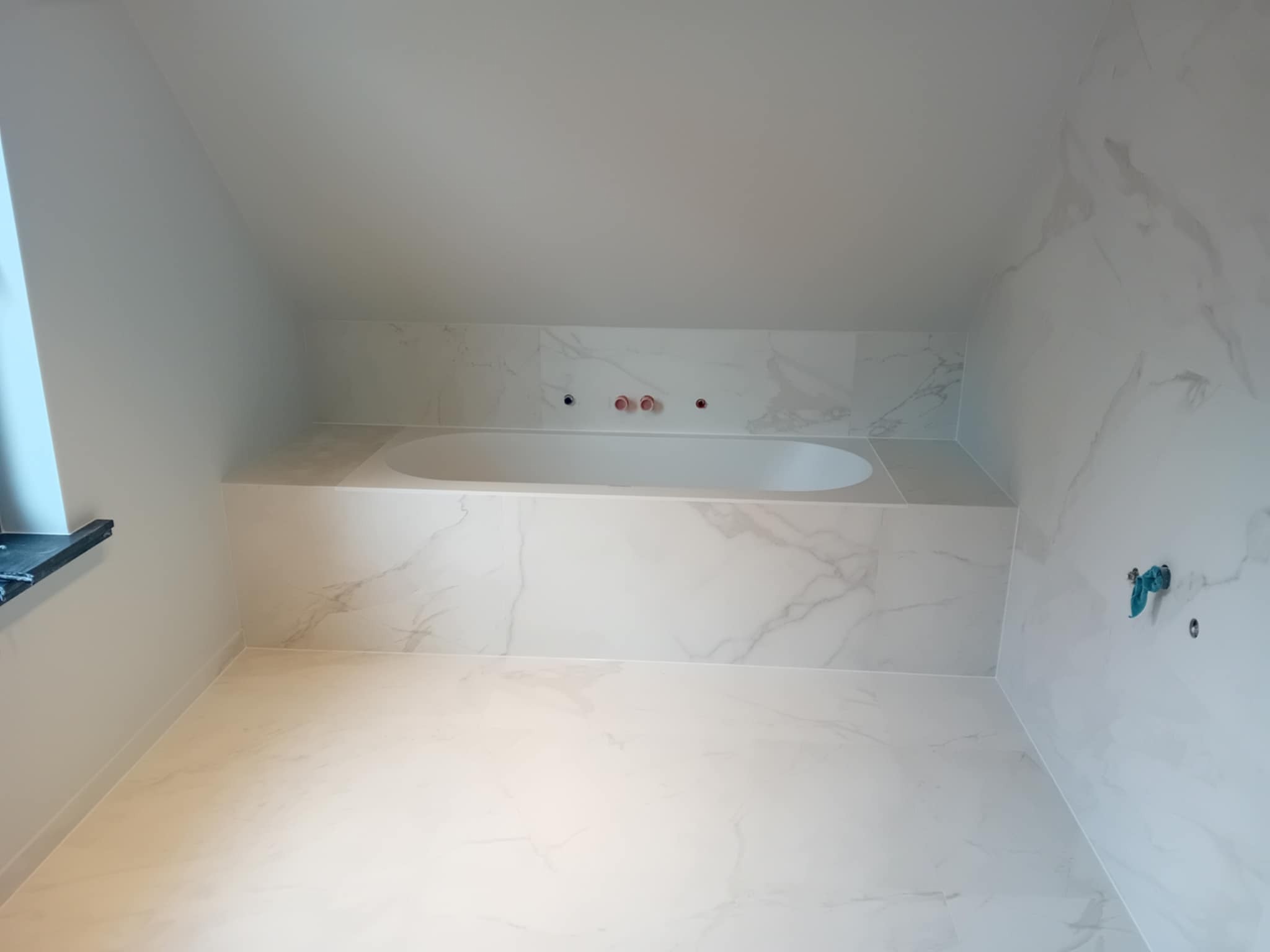 Wykończona łazienka na poddaszu z wanną obudowaną płytkami imitującymi marmur. Widoczne przyłącza wodne i okno. Minimalistyczny design z przewagą bieli i szarości.