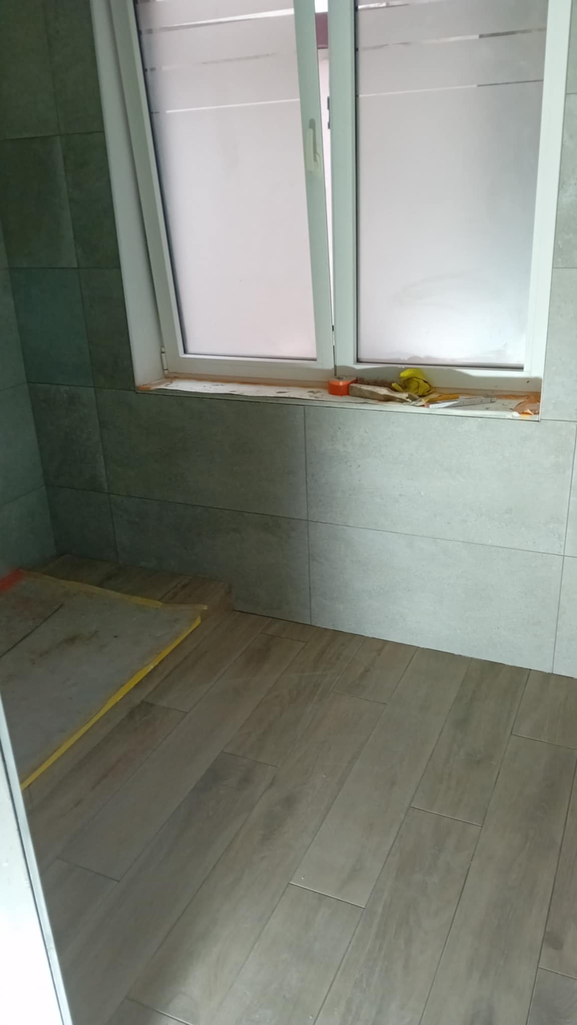Ułożone płytki podłogowe imitujące drewno w łazience z oknem. Widoczne narzędzia i materiały budowlane. Szare płytki ścienne tworzą spójną całość z podłogą.