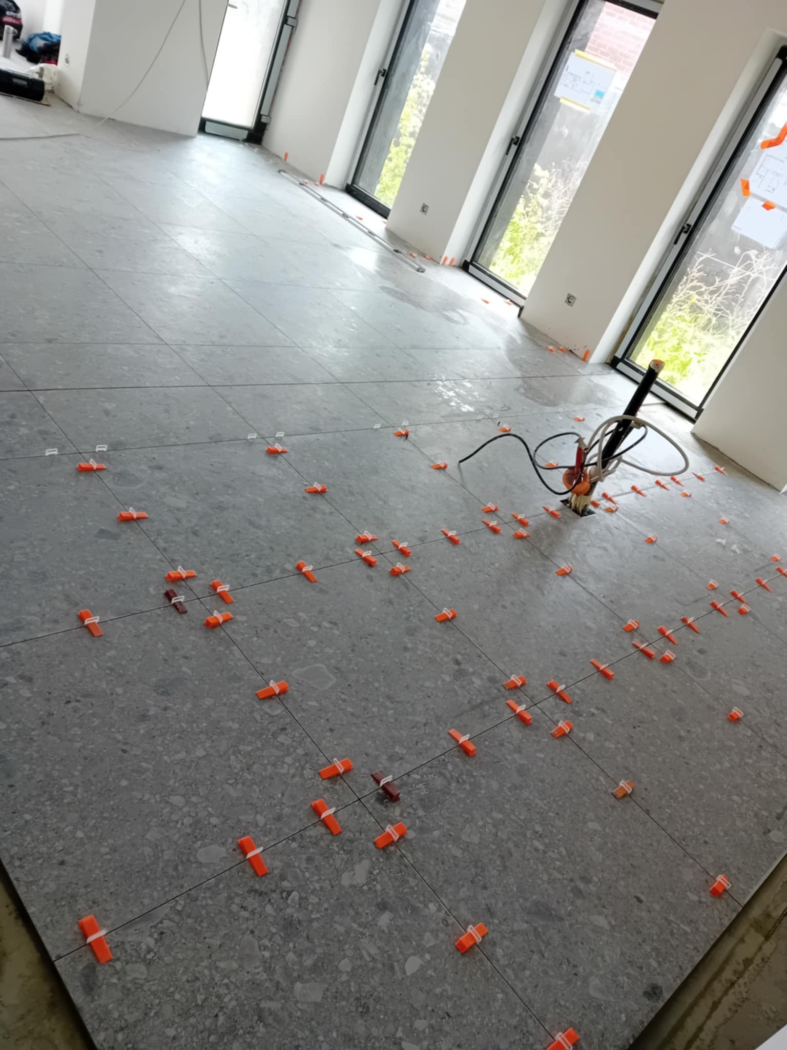 Ułożone szare płytki podłogowe z pomarańczowymi klipsami poziomującymi w przestronnym, jasnym pomieszczeniu z oknami. Widoczny sprzęt glazurniczy.