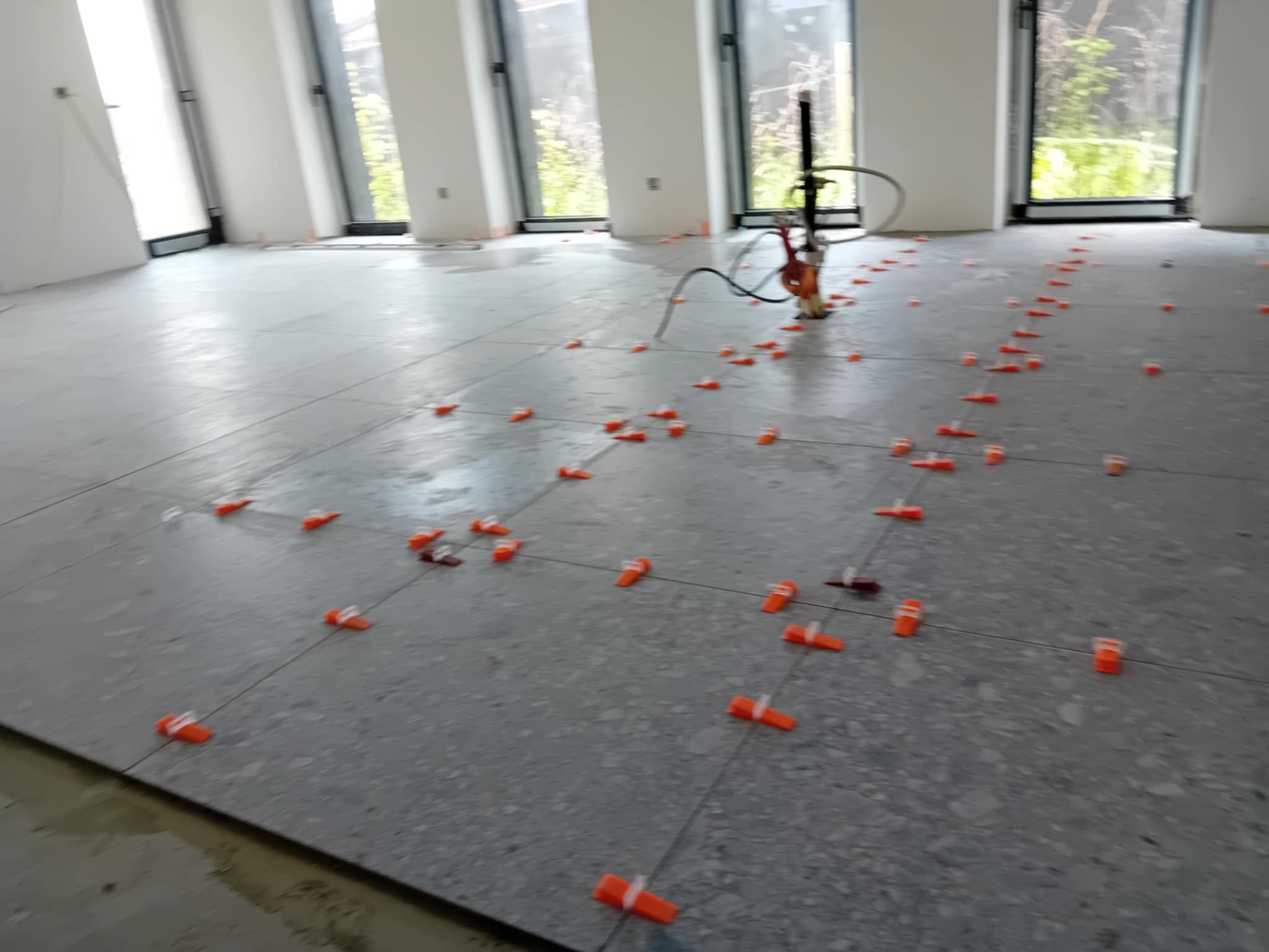 Szare płytki podłogowe w trakcie układania, z pomarańczowymi klipsami poziomującymi. W tle widoczne okna i narzędzie glazurnicze.