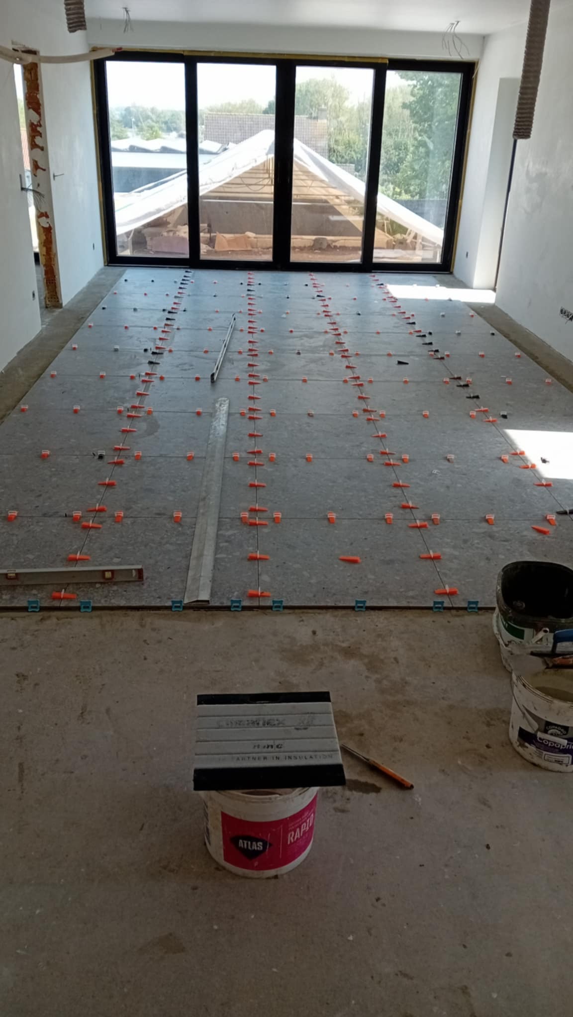 Układanie szarych płytek podłogowych z pomarańczowymi krzyżykami dystansowymi w jasnym pomieszczeniu z dużym oknem. Widoczne narzędzia i wiaderka z zaprawą.