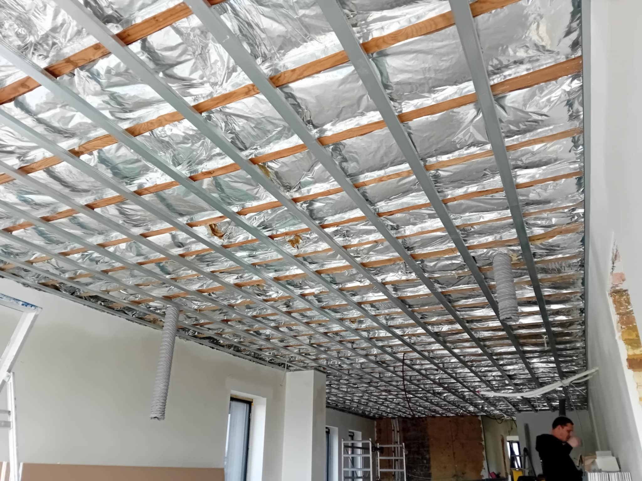 Sufit podwieszany w trakcie montażu: metalowa konstrukcja, izolacja folią aluminiową, widoczne drewniane elementy. Prace wykończeniowe w pomieszczeniu.