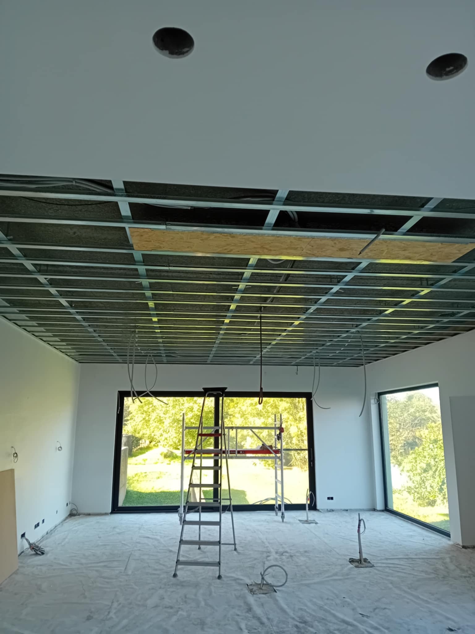 Sufit podwieszany w trakcie montażu: metalowa konstrukcja kratowa, częściowo zakryta płytami, widoczne przewody, drabina i zabezpieczona folią podłoga.