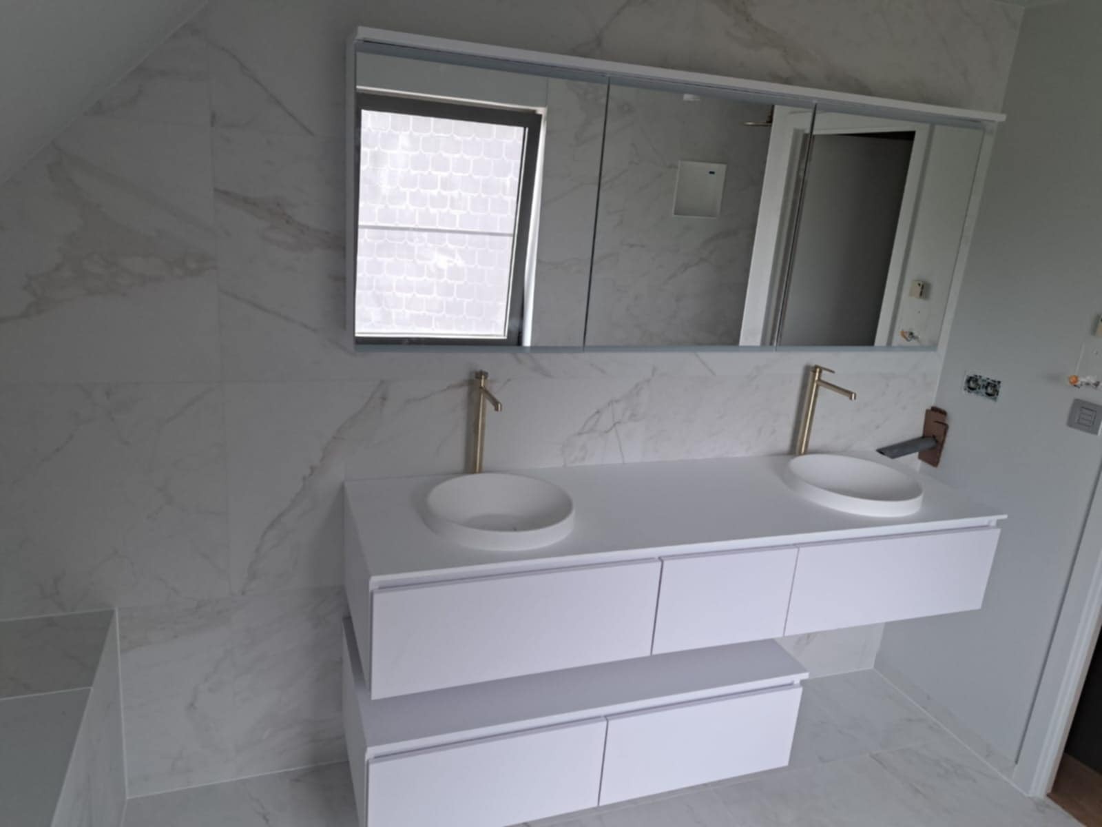 Wykończona łazienka w bieli: marmurowe płytki, podwójna umywalka z szafką i złotą armaturą, duże lustro z oknem. Nowoczesny design i dbałość o detale.
