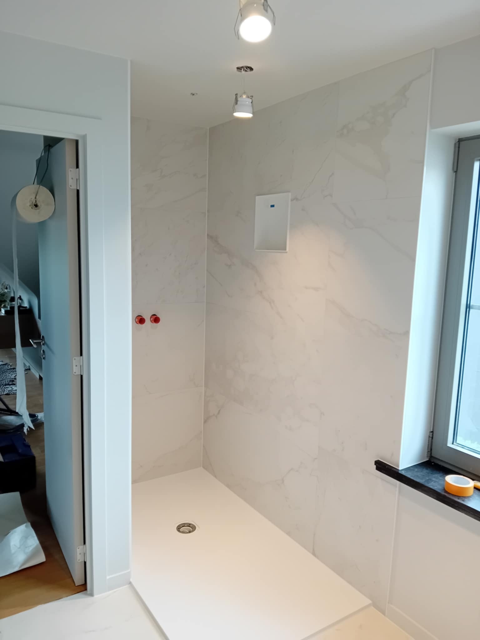 Nowoczesna kabina prysznicowa z białymi płytkami imitującymi marmur i odpływem liniowym. Widoczne okno i otwór na głośnik w ścianie. Minimalistyczny design.