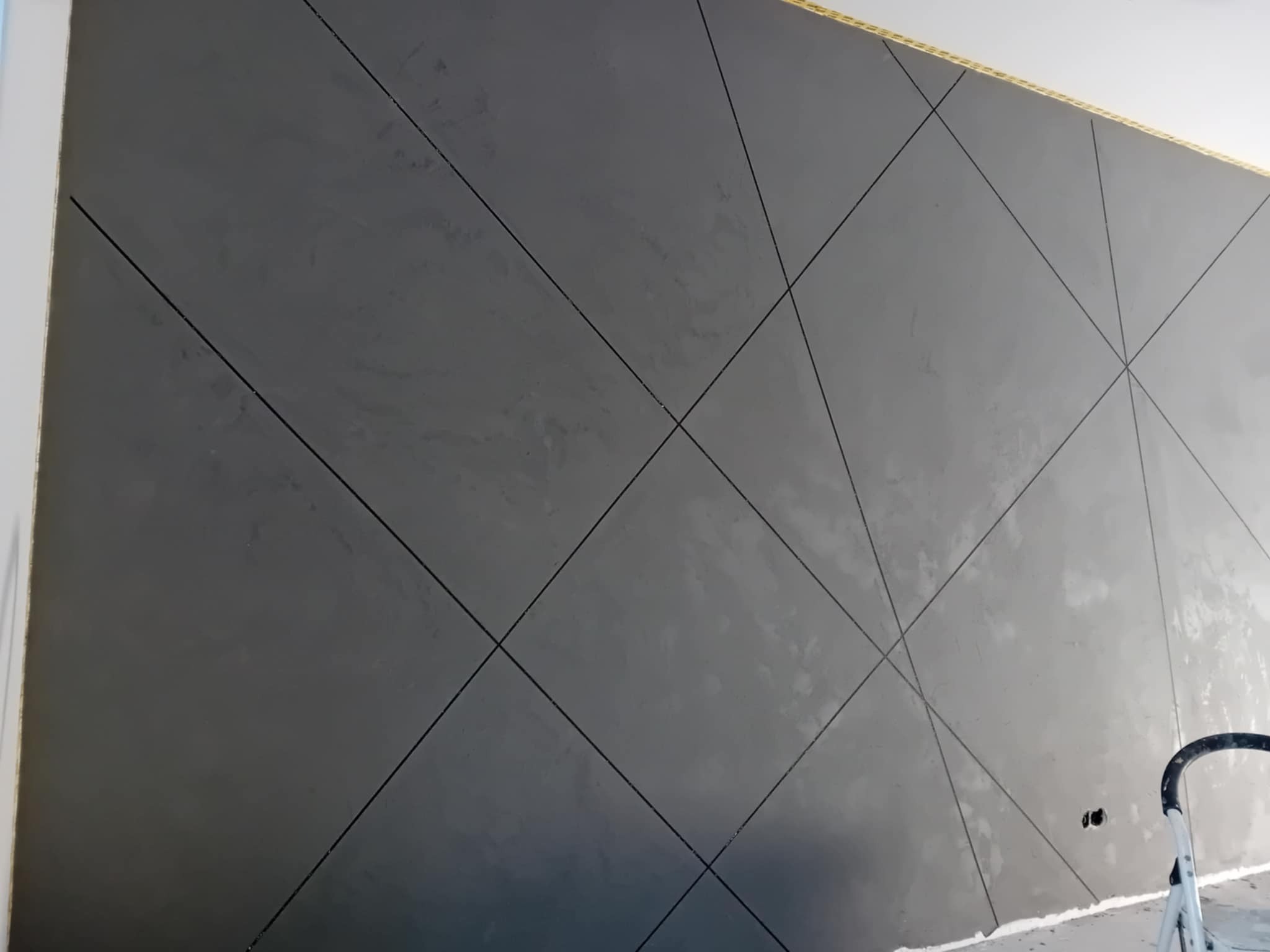 Szara ściana z geometrycznym wzorem w romby, uzyskana techniką dekoracyjną. Widoczne delikatne nierówności faktury i linie podziału. Fragment drabiny w prawym dolnym rogu.