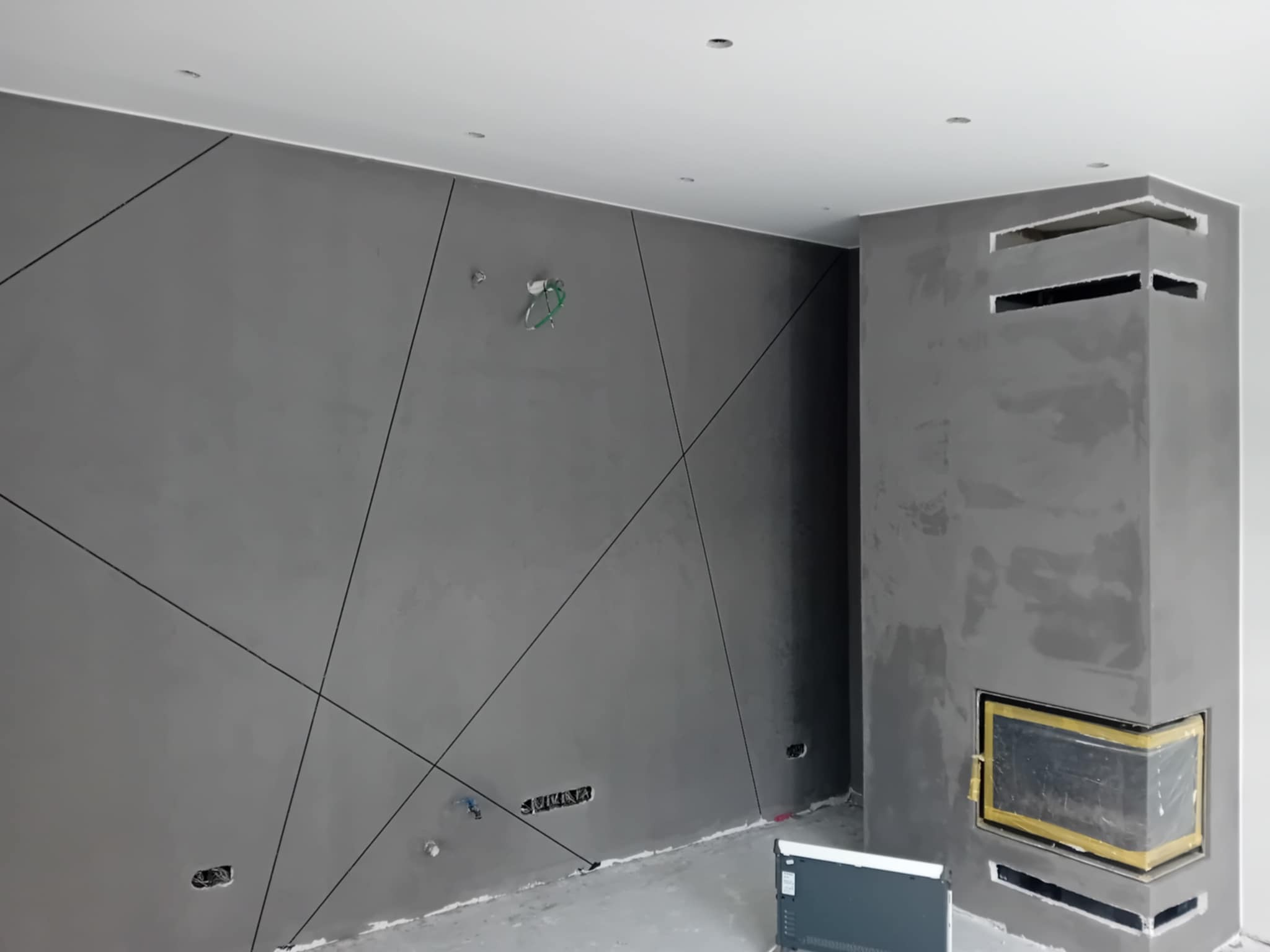 Szara ściana z geometrycznym wzorem i kominek w surowym wykończeniu. Widoczne niedokończone instalacje elektryczne. Podłoga w stanie surowym.