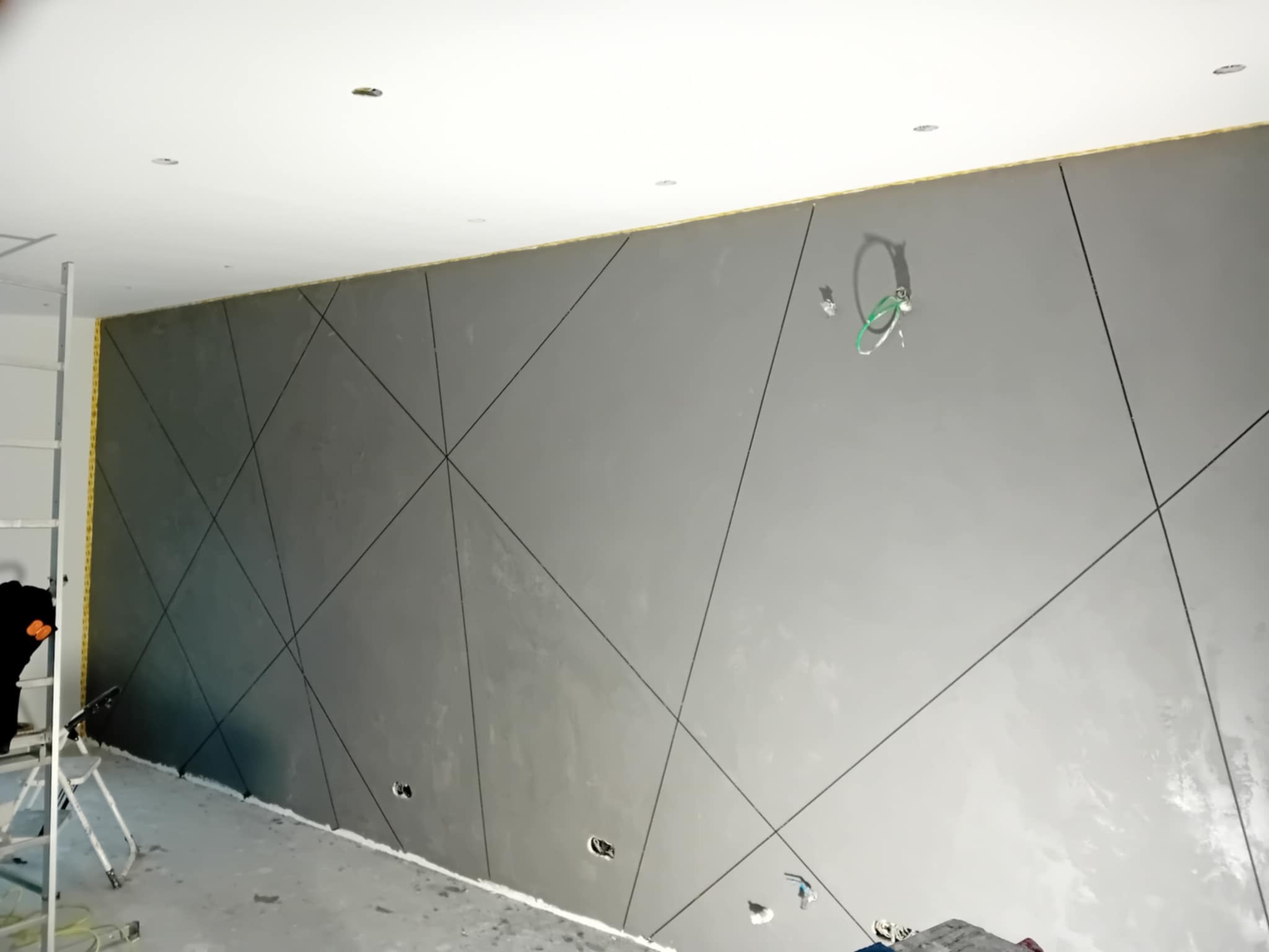 Szara ściana z geometrycznym wzorem w romby i trójkąty, widoczne puszki elektryczne i drabina, świeżo wykończone wnętrze w trakcie prac wykończeniowych.