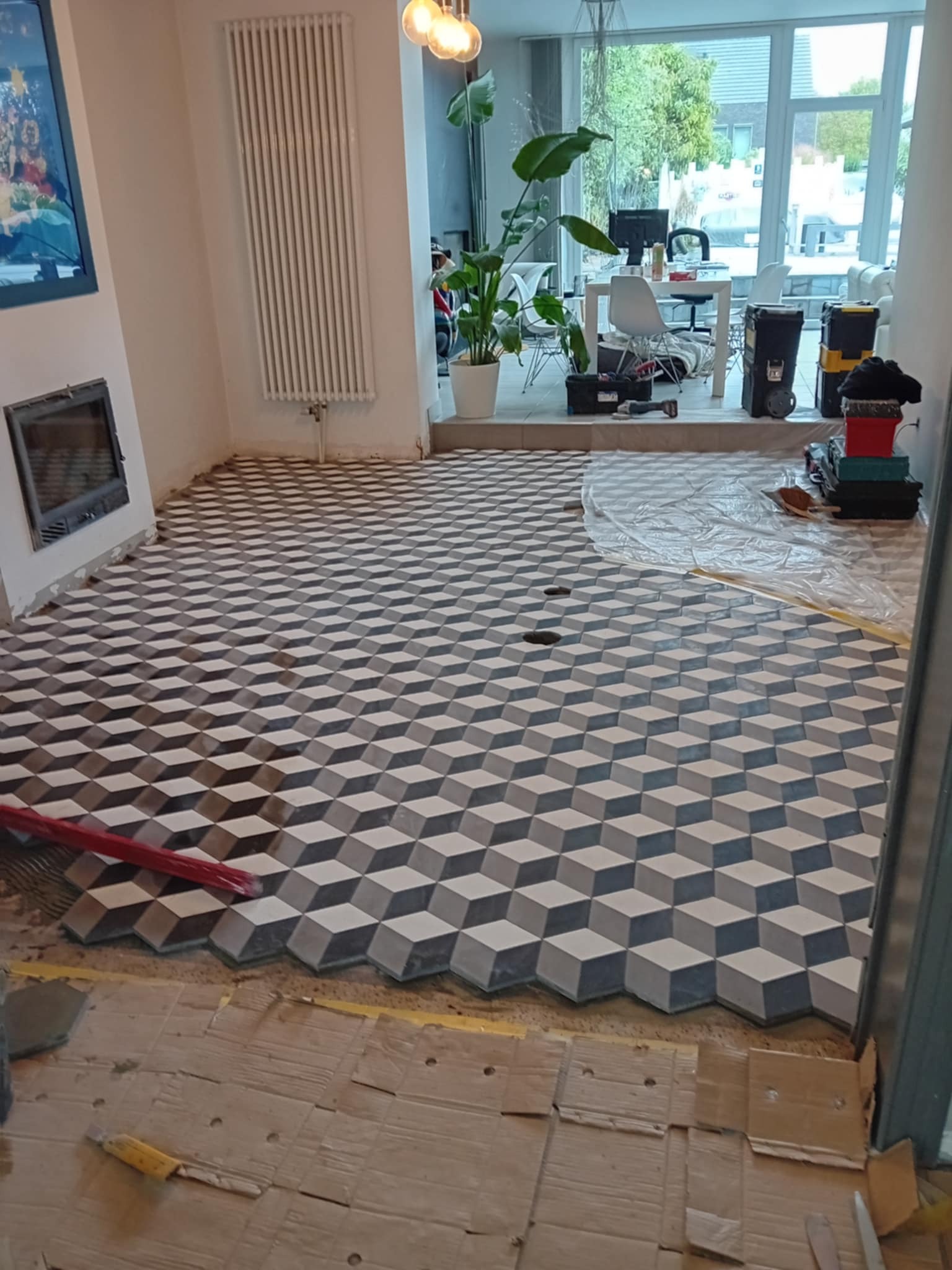 Ukończona podłoga z geometrycznych płytek 3D w odcieniach szarości, z widocznymi narzędziami i elementami w trakcie montażu, w jasnym, przestronnym wnętrzu.