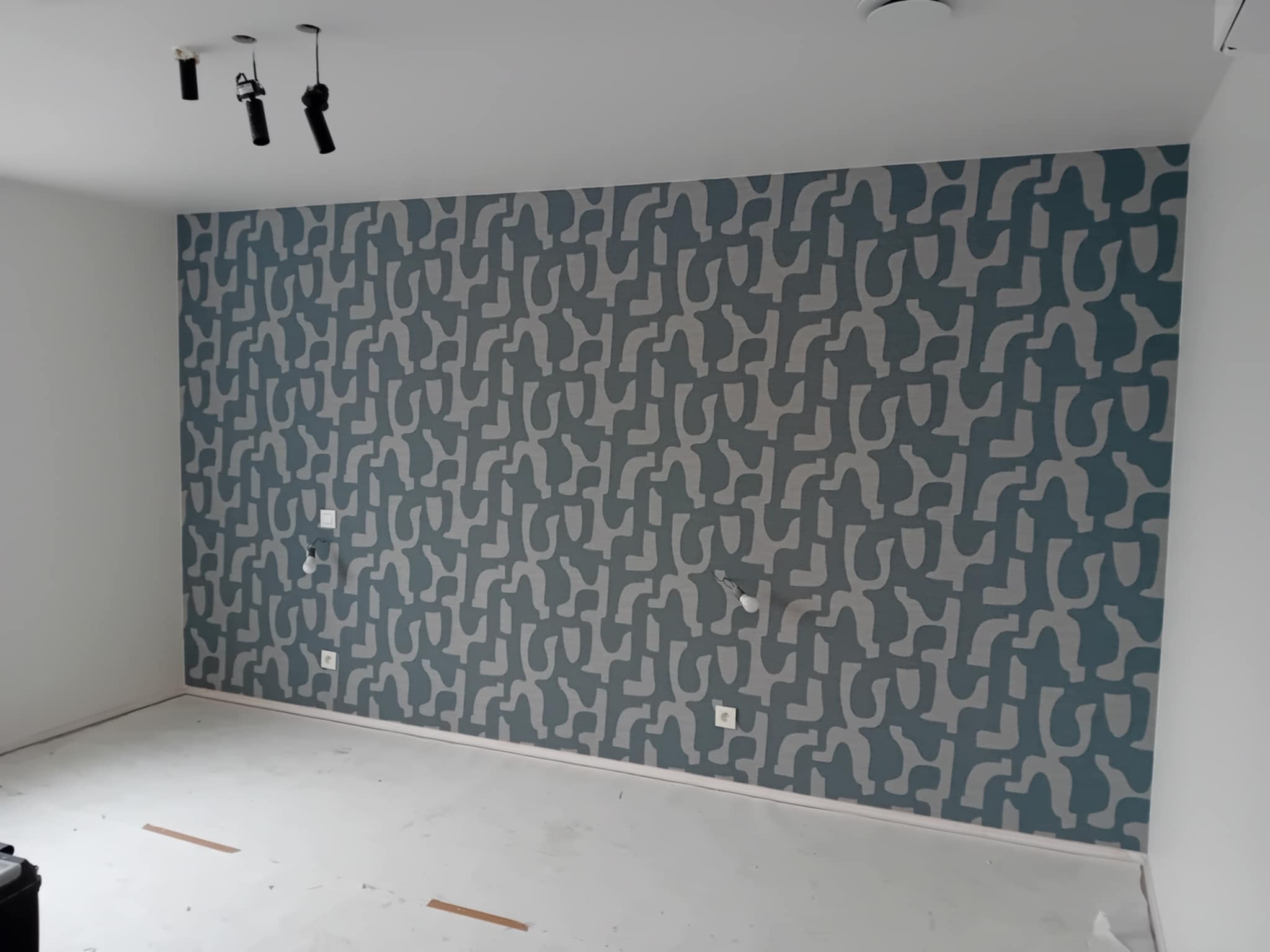 Ściana z wzorzystą tapetą w odcieniach szarości i błękitu, widoczne gniazdka elektryczne i listwy przypodłogowe, sufit z reflektorami punktowymi, surowe wykończenie podłogi.