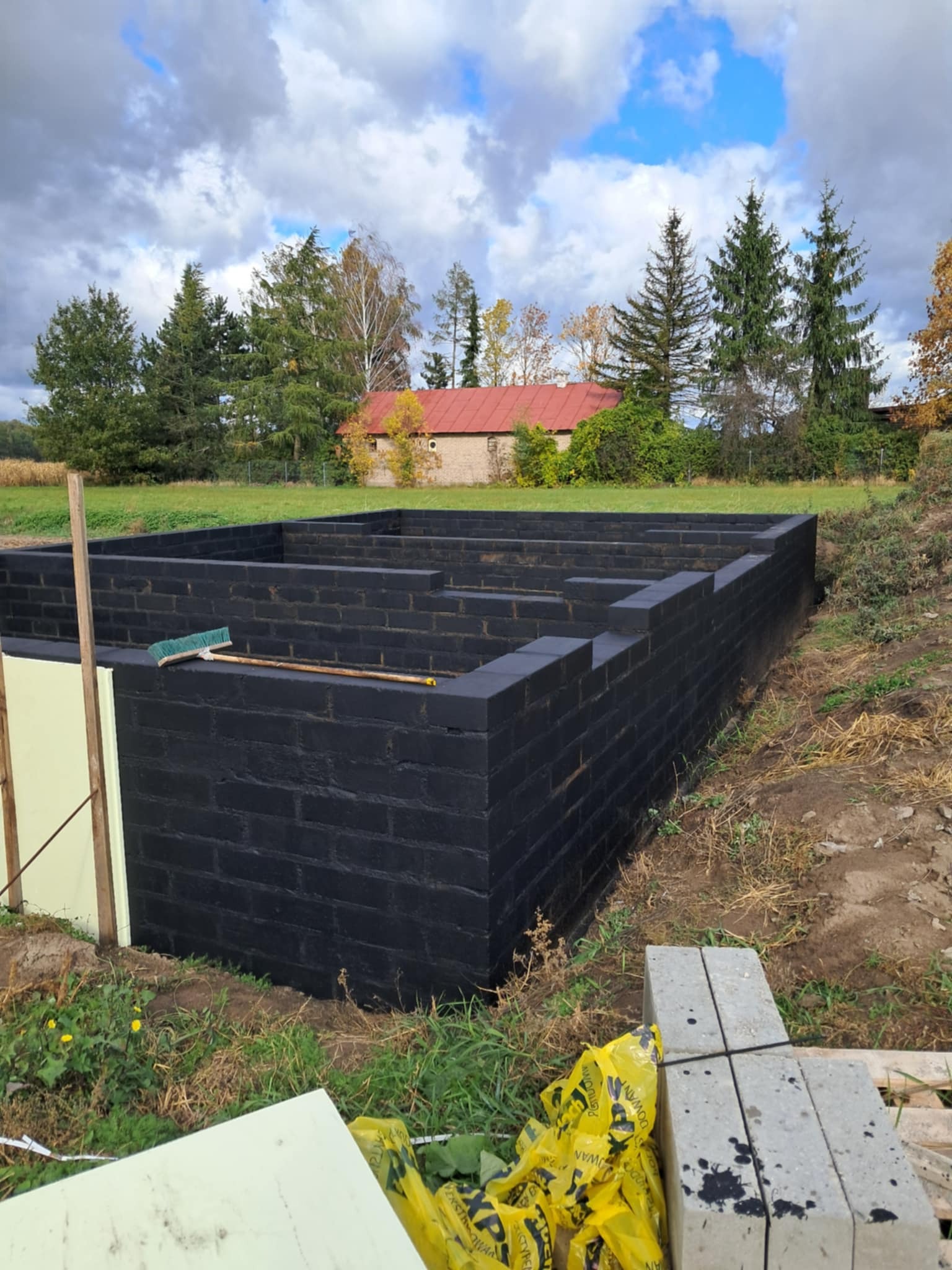 Fundamenty z czarnej cegły na zewnątrz, z widoczną izolacją i szczotką na murze. Tło stanowią drzewa i budynek z czerwonym dachem.