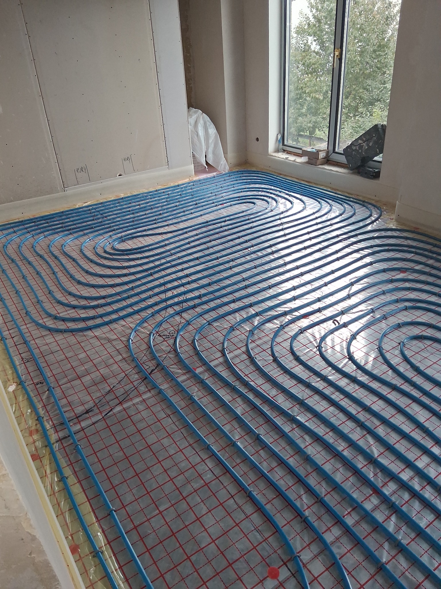 Niebieskie rury ogrzewania podłogowego ułożone na folii z czerwoną kratką w jasnym pomieszczeniu z oknem. Widoczne płyty kartonowo-gipsowe na ścianie.