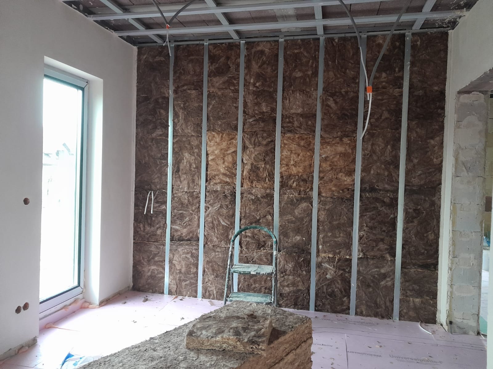 Ocieplenie ścian wełną mineralną w trakcie montażu, widoczne profile metalowe. Na podłodze różowa folia. Drabina oparta o ścianę. Widok na okno i surowy stan pomieszczenia.