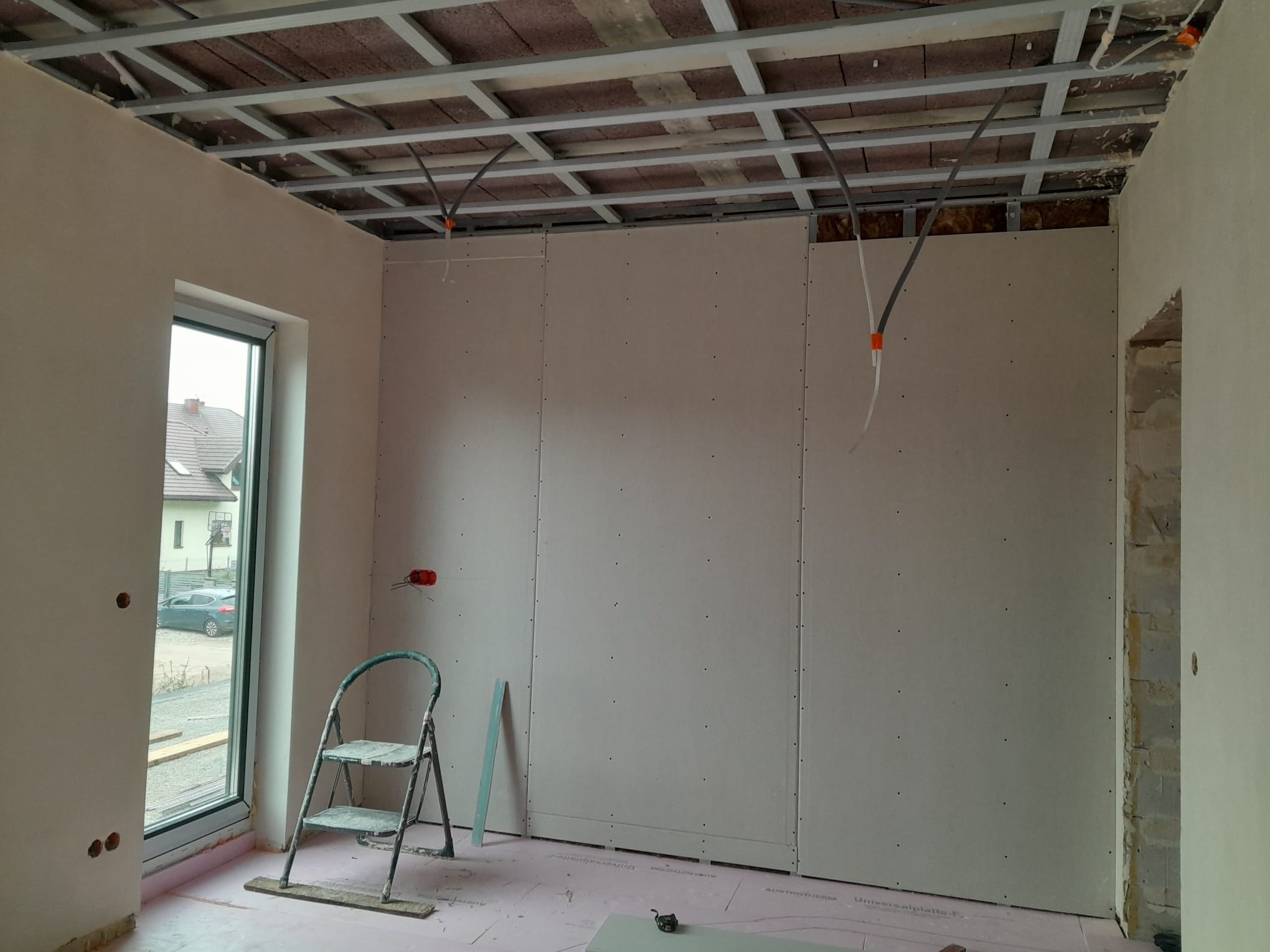 Wnętrze w trakcie montażu płyt karton-gips na ścianach i suficie. Widoczne profile, instalacje elektryczne i drabina. Podłoga zabezpieczona folią. Okno z widokiem na dom.