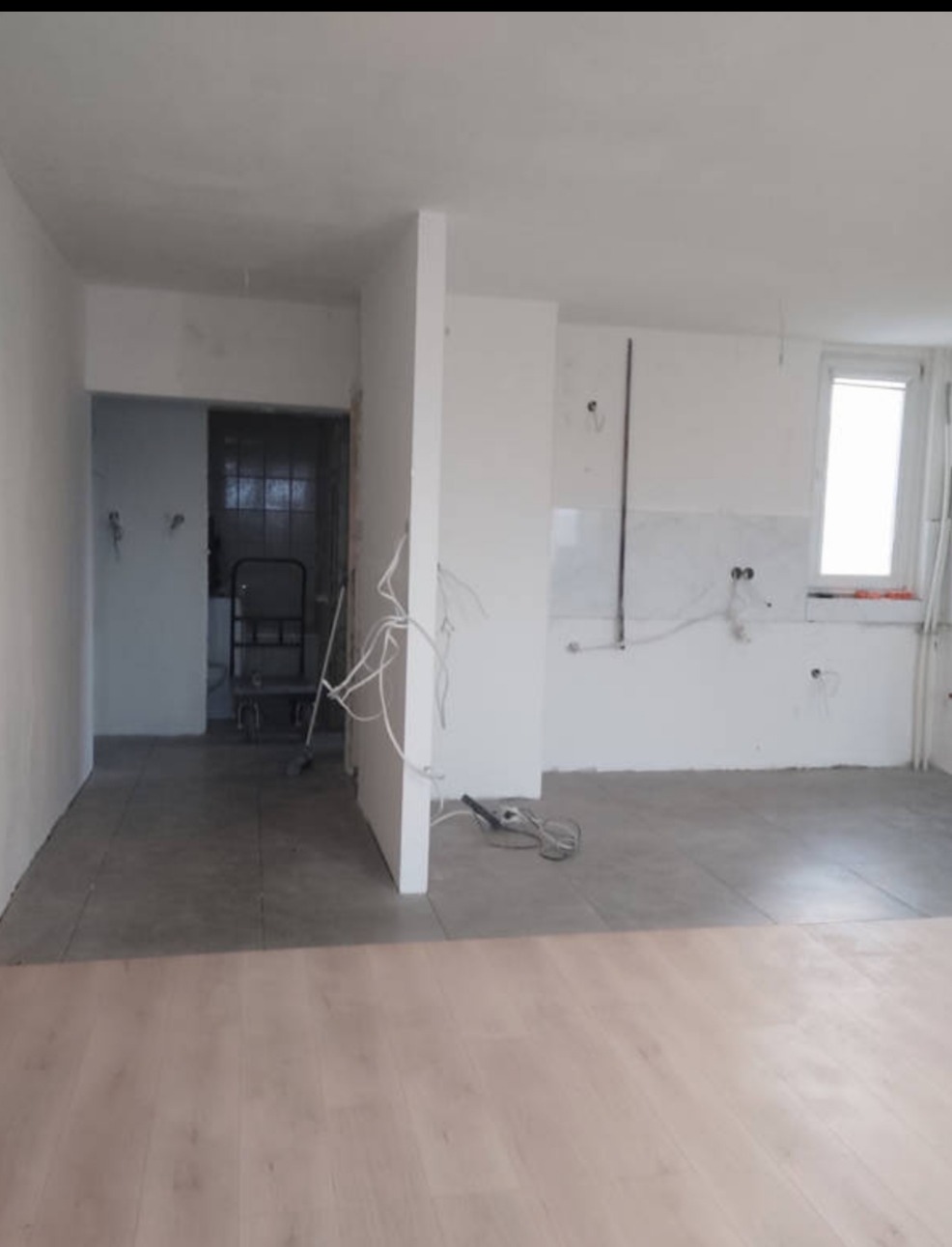 Wnętrze w trakcie remontu: szare płytki podłogowe, białe ściany z widoczną instalacją elektryczną, oraz fragment nowej, jasnej podłogi. Widoczny wózek transportowy.