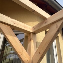 PRACOWNIA STOLARSKA DOMINIK PIONTEK - Fragment konstrukcji drewnianej, prawdopodobnie zadaszenia tarasu, z widocznymi elementami mocującymi i fakturą drewna na tle elewacji budynku.