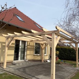 PRACOWNIA STOLARSKA DOMINIK PIONTEK - Surowa, drewniana konstrukcja pergoli na tarasie z kostki brukowej, z widokiem na dom z czerwoną dachówką i zielony żywopłot w tle. Wczesna faza budowy.