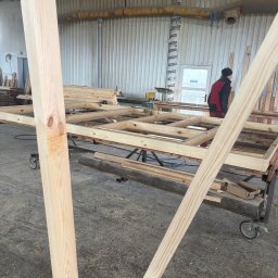 PRACOWNIA STOLARSKA DOMINIK PIONTEK - Wnętrze warsztatu: drewniana konstrukcja szkieletowa na wózku, przygotowywana do dalszej obróbki. W tle widoczna osoba i elementy wyposażenia stolarni.