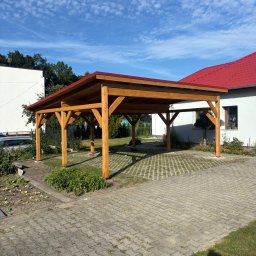 PRACOWNIA STOLARSKA DOMINIK PIONTEK - Drewniana wiata garażowa z czerwonym dachem na posesji z kostką brukową i zielenią. Konstrukcja z belek, widok z zewnątrz, słoneczny dzień.
