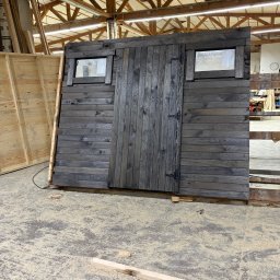 PRACOWNIA STOLARSKA DOMINIK PIONTEK - Ciemna, drewniana konstrukcja z drzwiami i oknami, prawdopodobnie fragment większej budowli, w warsztacie. Widoczne ślady obróbki drewna na podłodze.