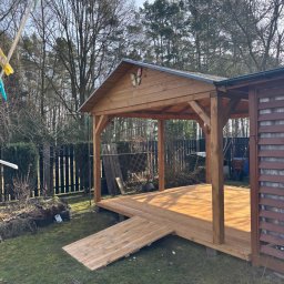 PRACOWNIA STOLARSKA DOMINIK PIONTEK - Drewniana altana z rampą wejściową, ozdobiona motylem na dachu, na tle ogrodu z drzewami i czarnym płotem. Słoneczny dzień.