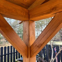 PRACOWNIA STOLARSKA DOMINIK PIONTEK - Detal drewnianej konstrukcji altany z widocznym słupem, belkami i fragmentem dachu. W tle zieleń i czarne ogrodzenie.