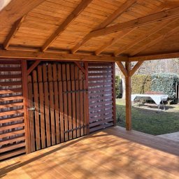 PRACOWNIA STOLARSKA DOMINIK PIONTEK - Zadaszona, drewniana konstrukcja z podłogą i ażurowymi ścianami, zamykana na drewniane wrota. W tle ogród z białym stołem i krzesłami.