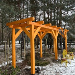 PRACOWNIA STOLARSKA DOMINIK PIONTEK - Nowa, pomarańczowa pergola w zimowym ogrodzie, z żółtą ławką i latarnią w tle, otoczona drzewami i śniegiem. Widok na konstrukcję w naturalnym otoczeniu.