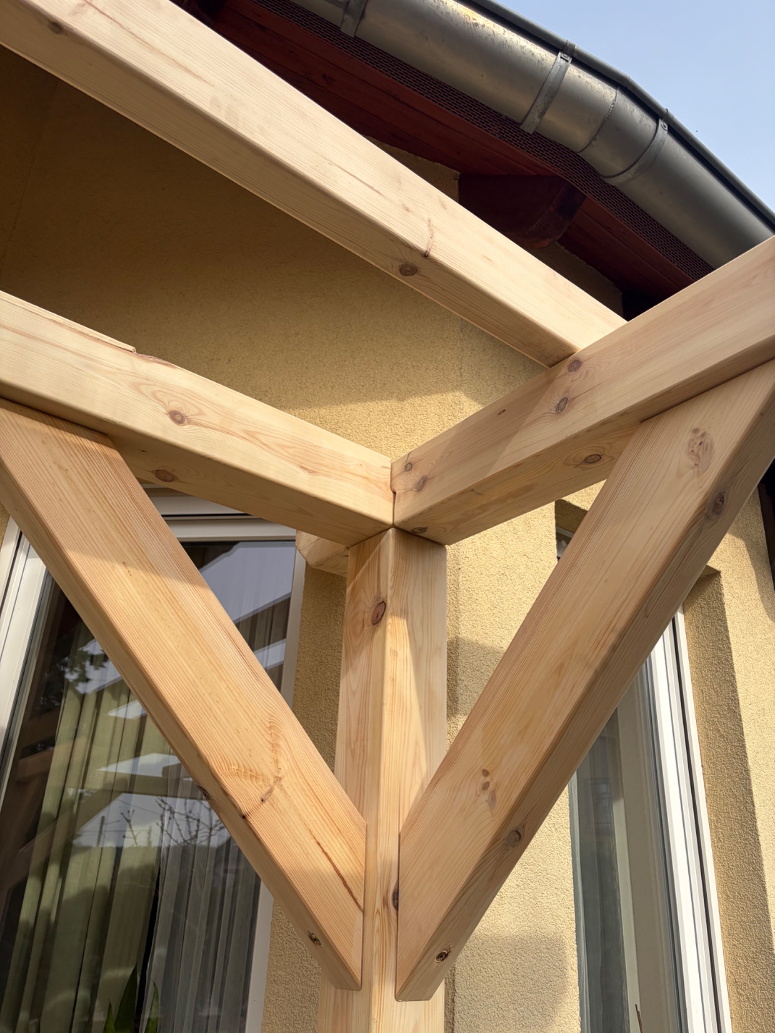 Fragment konstrukcji drewnianej, prawdopodobnie zadaszenia tarasu, z widocznymi elementami mocującymi i fakturą drewna na tle elewacji budynku.