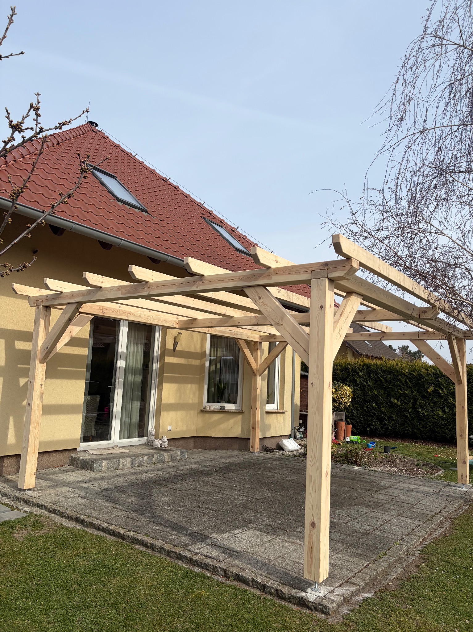 Surowa, drewniana konstrukcja pergoli na tarasie z kostki brukowej, z widokiem na dom z czerwoną dachówką i zielony żywopłot w tle. Wczesna faza budowy.