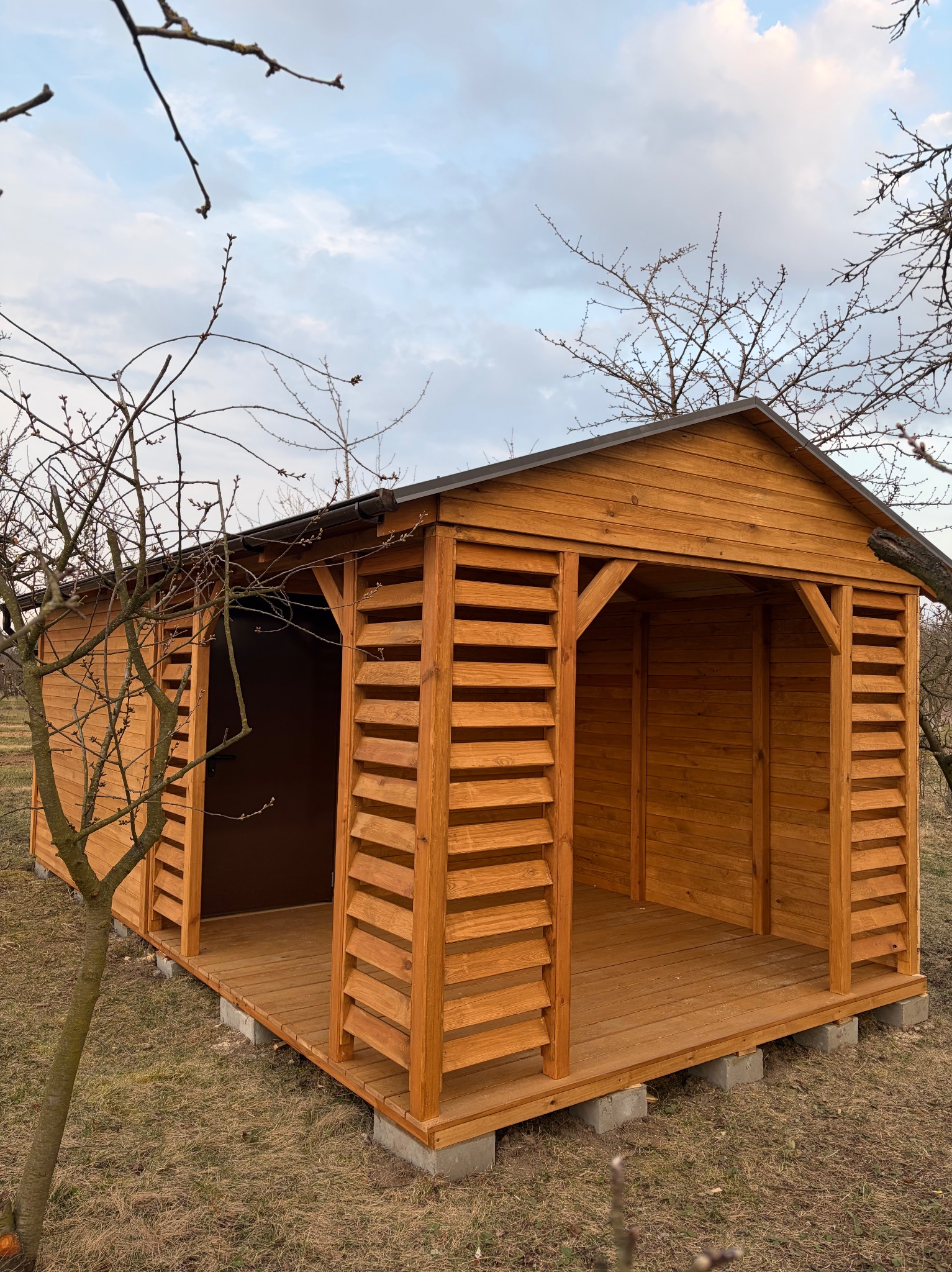 Drewniana altana ogrodowa z lamelowymi ścianami, osadzona na betonowych bloczkach, z widoczną konstrukcją dachu i podłogą z desek. Ujęcie wczesną wiosną.