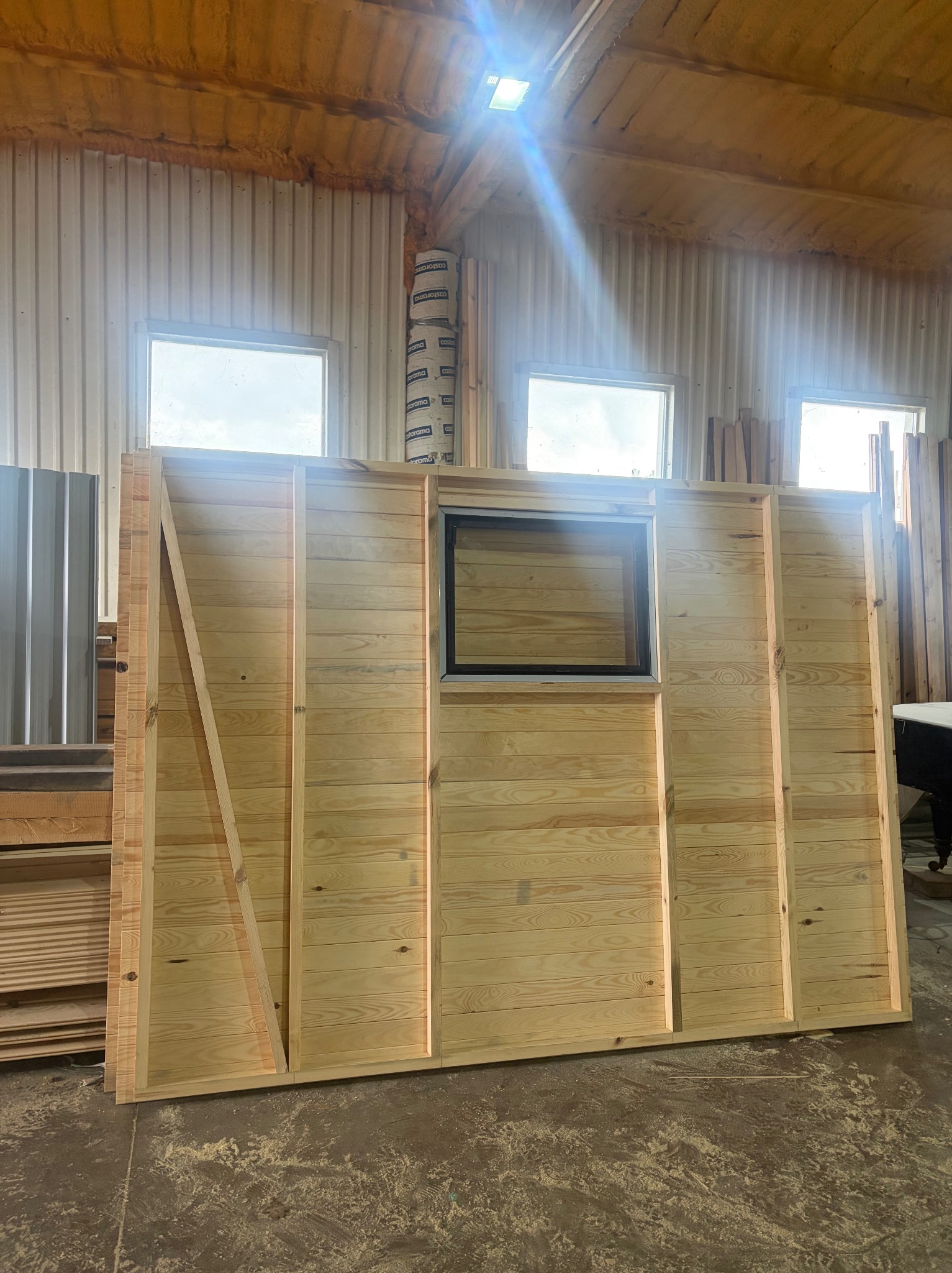 Element drewnianej konstrukcji z oknem, prawdopodobnie fragment altany lub domku narzędziowego, w warsztacie z widocznymi deskami i elementami konstrukcyjnymi.