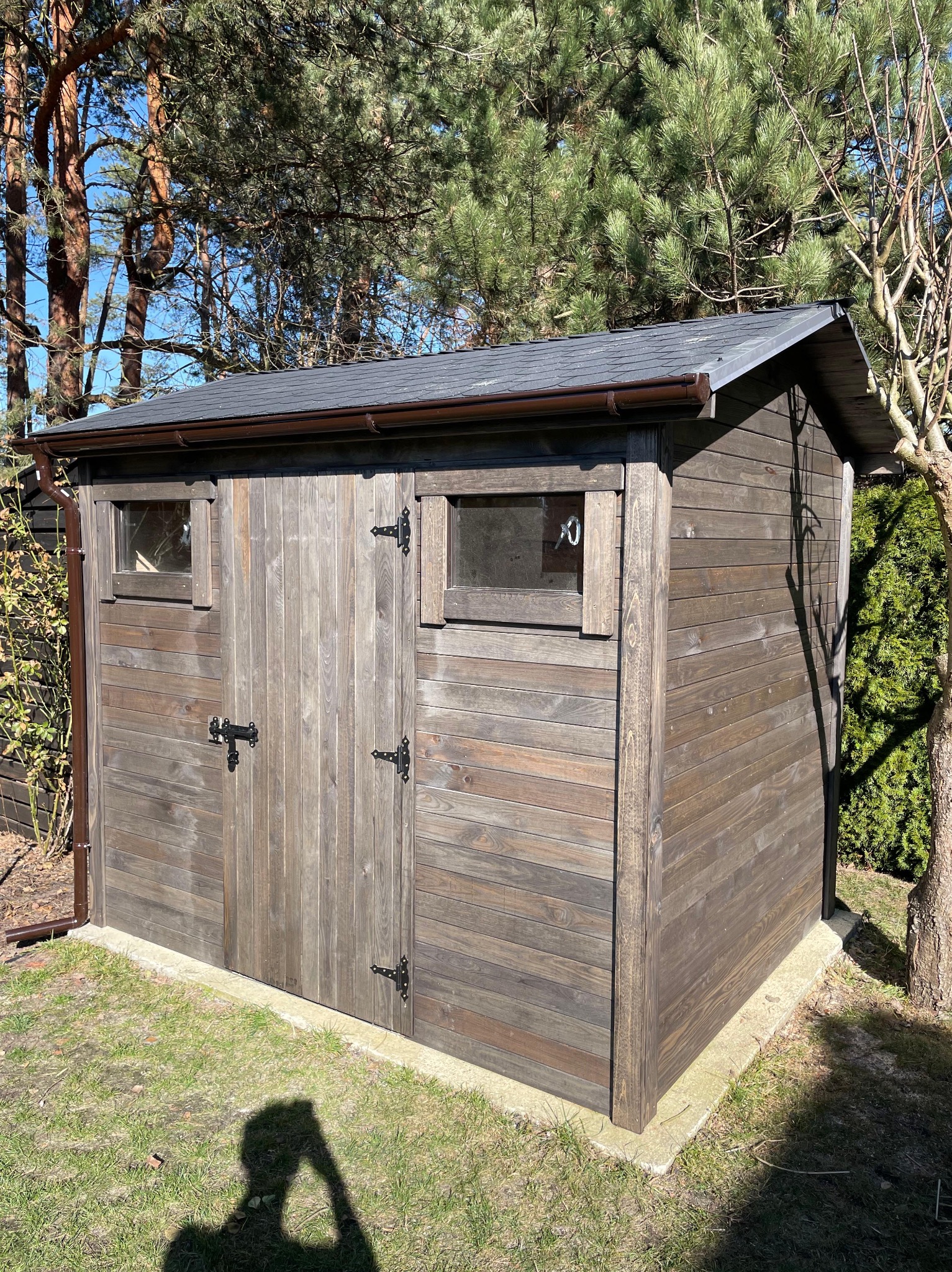 Drewniana szopa narzędziowa z dwuspadowym dachem, brązowymi rynnami i małymi oknami, otoczona zielenią drzew i trawnikiem w słoneczny dzień. Widoczny cień osoby.
