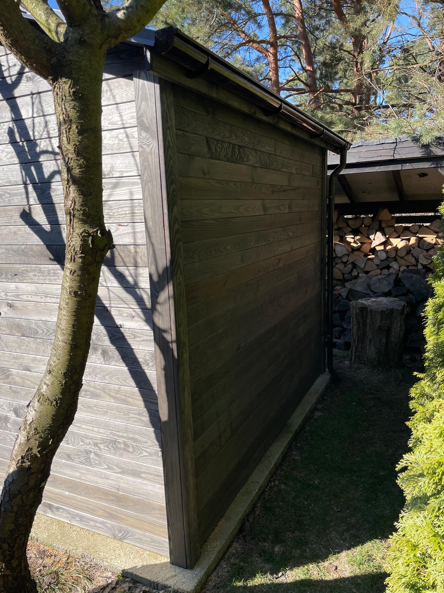 Drewniana altana z ciemnego drewna, widoczna z boku, z pniem drzewa w kadrze. W tle skład drewna pod zadaszeniem i iglaki. Słoneczny dzień.