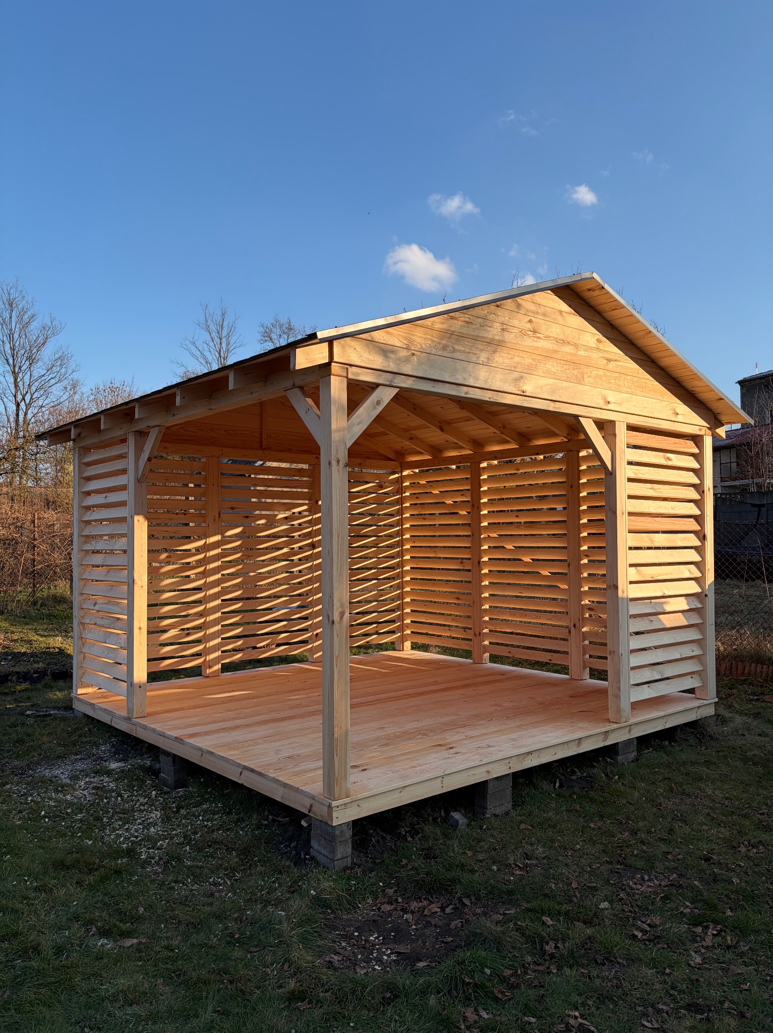Surowa, drewniana altana z lamelami, wzniesiona na betonowych bloczkach, na tle trawnika i błękitnego nieba. Widoczne detale konstrukcyjne i naturalne oświetlenie.
