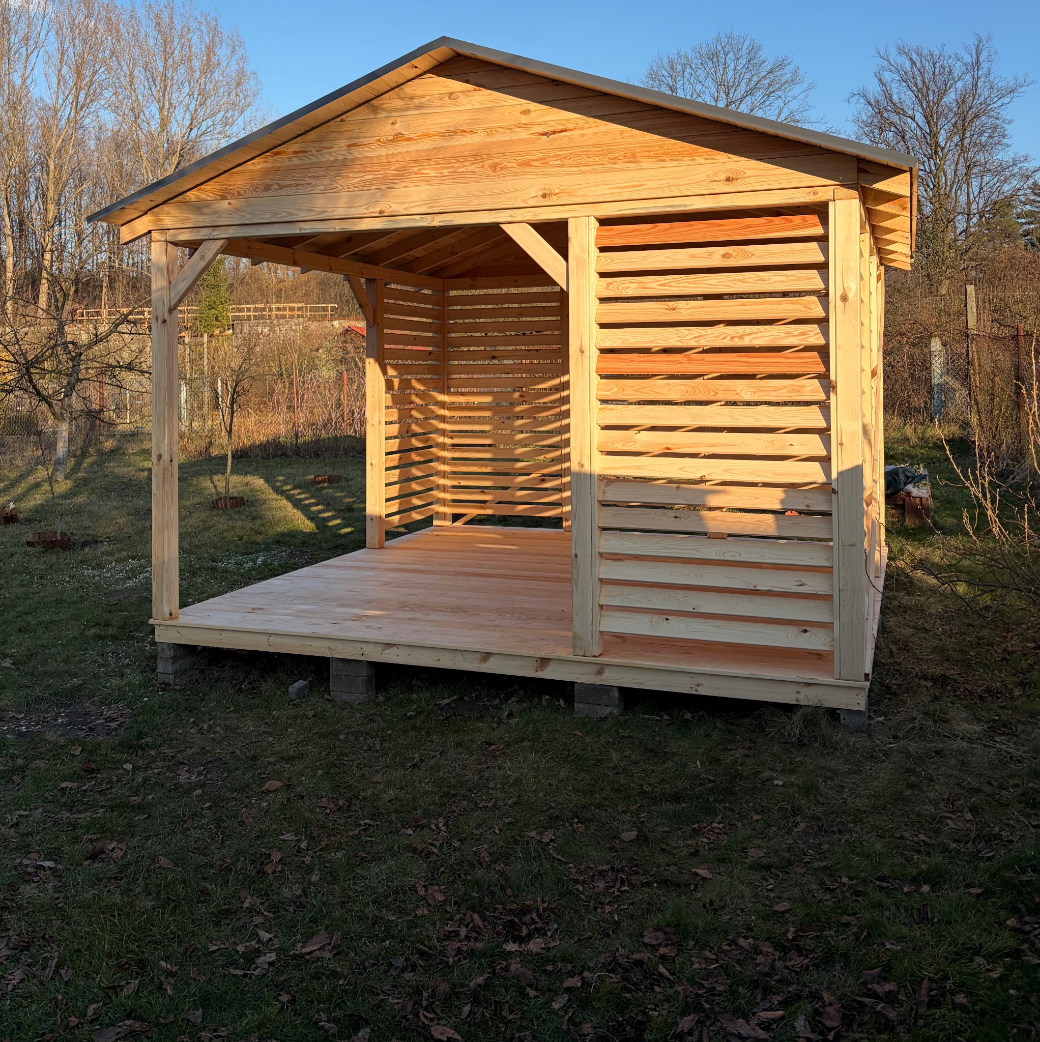 Surowa, drewniana konstrukcja altany z lamelami na ścianach bocznych, osadzona na betonowych bloczkach, na tle ogrodu w jesiennym słońcu.