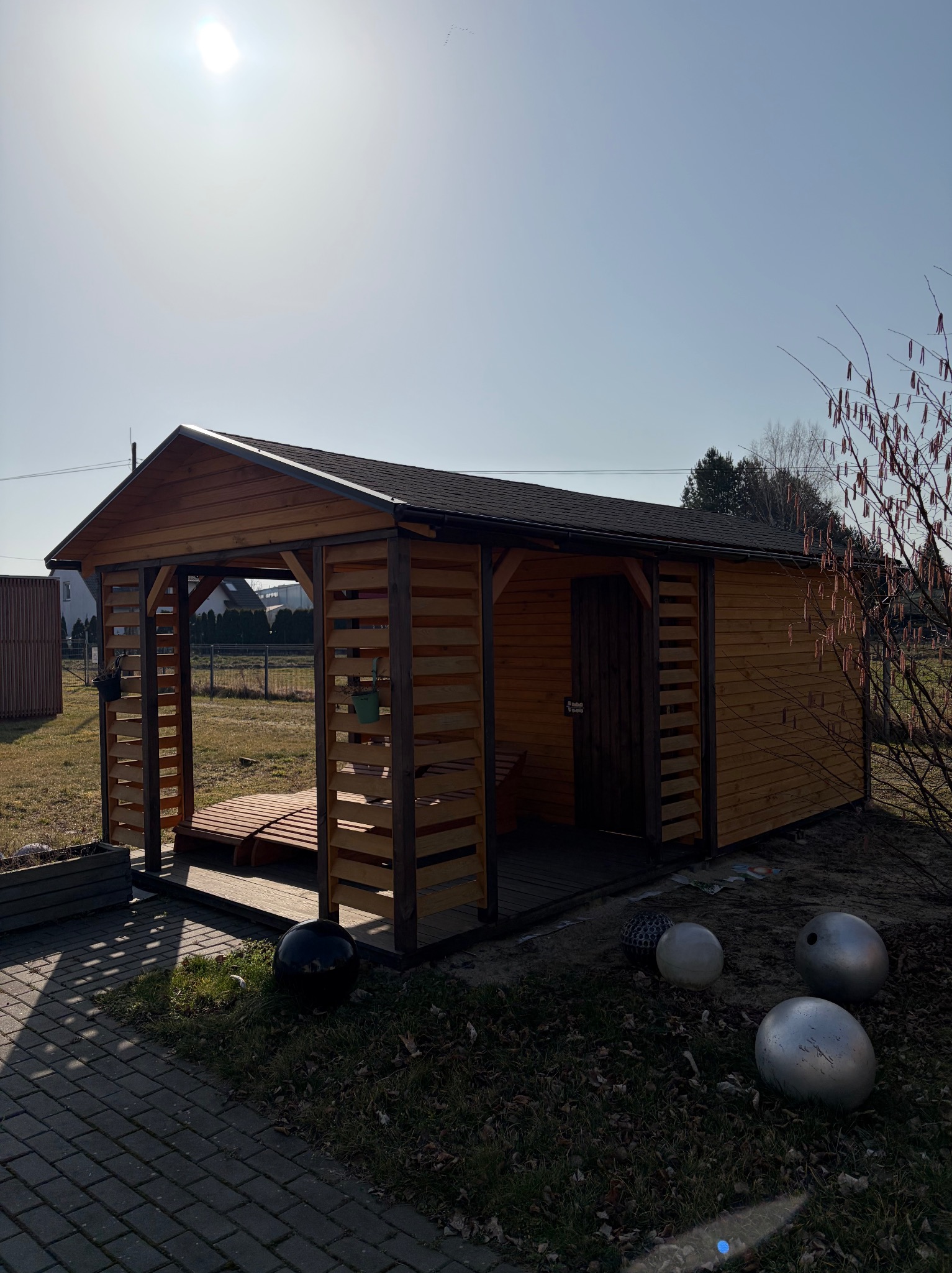 Drewniana altana z zadaszeniem, częściowo zabudowana, z drewnianą podłogą i dekoracyjnymi kulami w otoczeniu. Widok w słoneczny dzień.