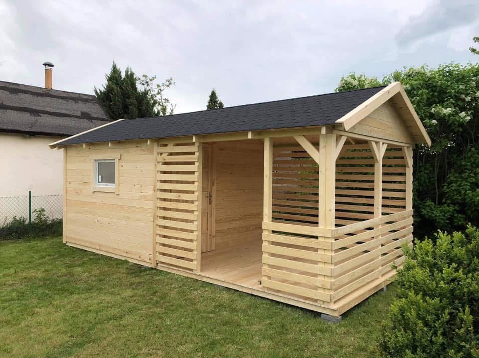 Surowa, drewniana konstrukcja z czarnym dachem, łącząca zamkniętą budę z otwartą przestrzenią z poziomymi szczeblami, na tle zielonego trawnika i zabudowań.