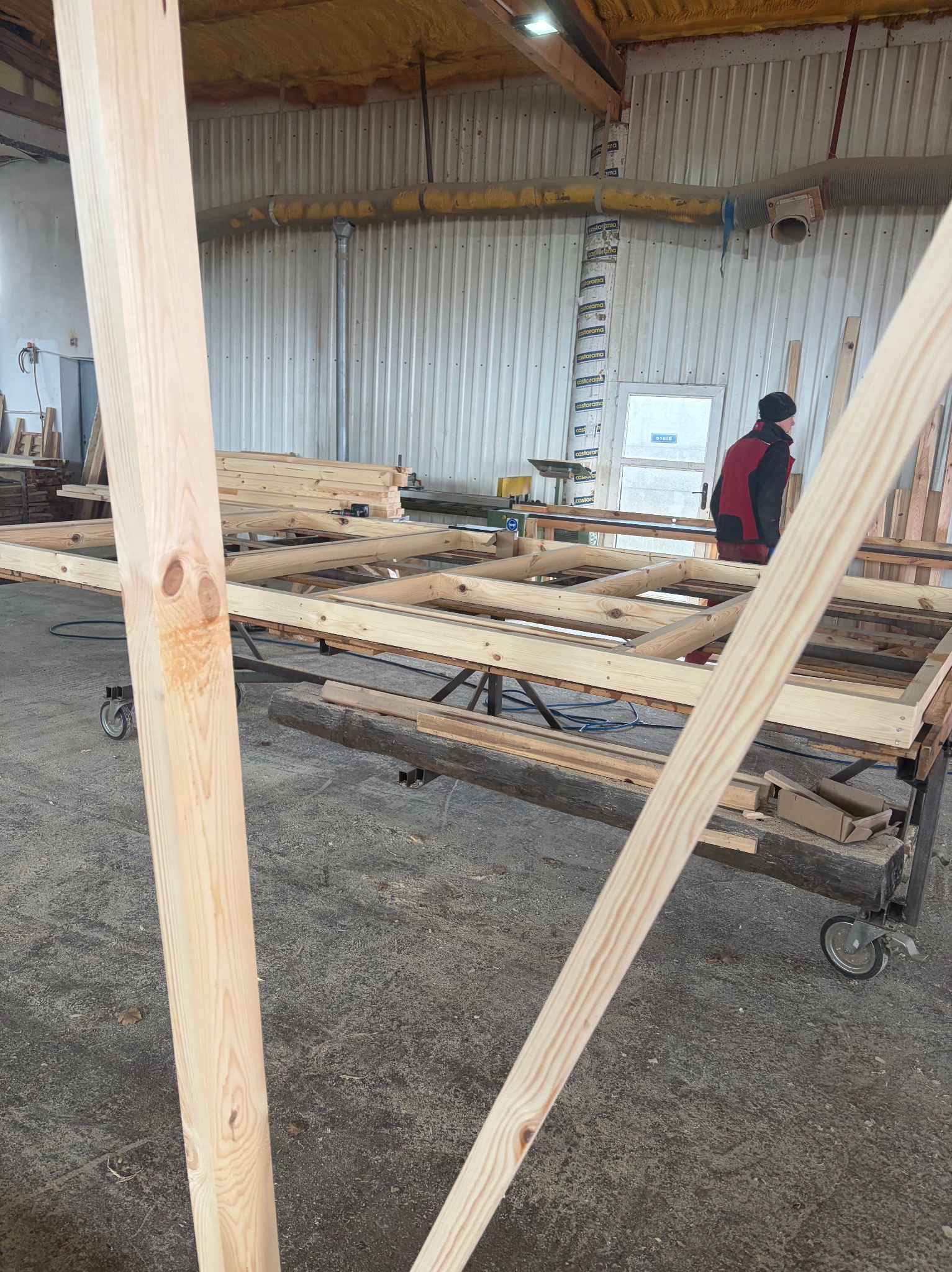 Wnętrze warsztatu: drewniana konstrukcja szkieletowa na wózku, przygotowywana do dalszej obróbki. W tle widoczna osoba i elementy wyposażenia stolarni.