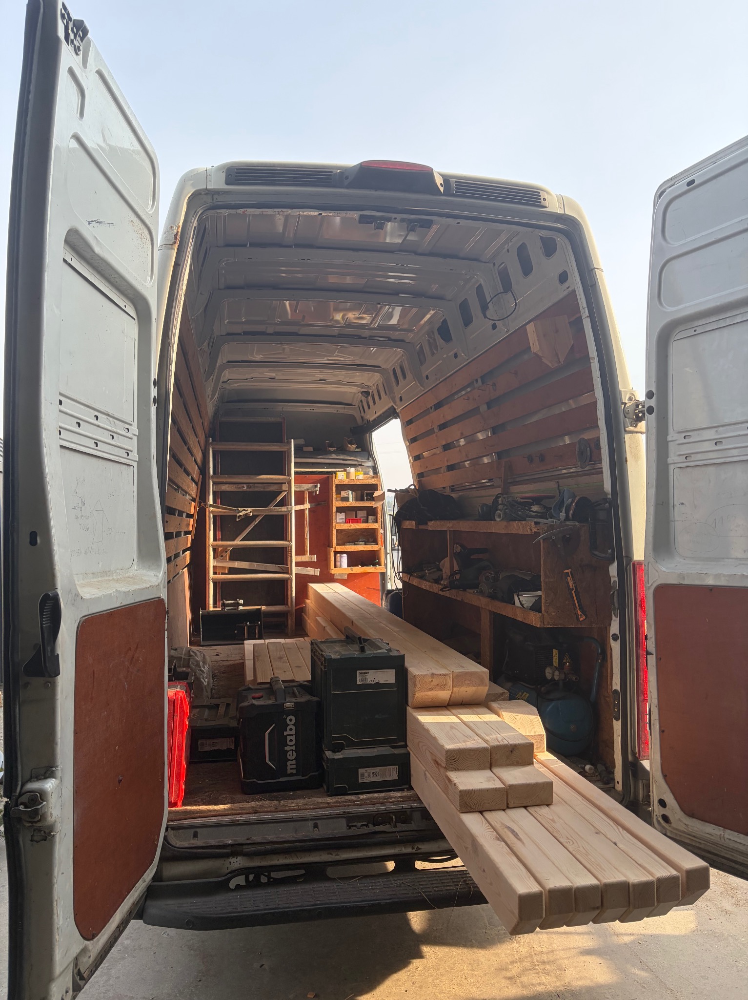 Wnętrze furgonetki z otwartymi drzwiami, wypełnione drewnem, narzędziami i skrzynkami Metabo. Widoczne półki i drabina. Przygotowanie do pracy stolarskiej.