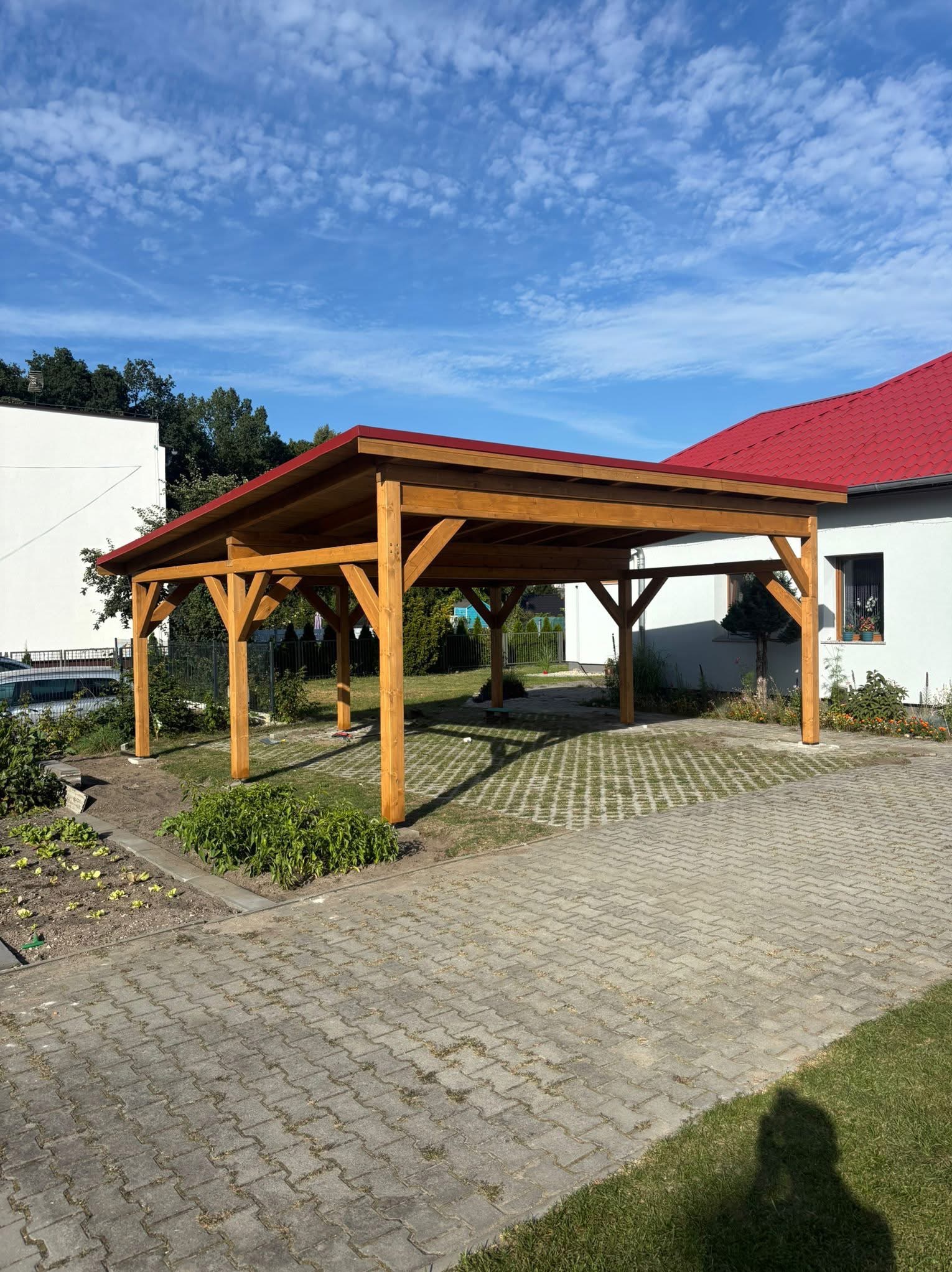 Drewniana wiata garażowa z czerwonym dachem na posesji z kostką brukową i zielenią. Konstrukcja z belek, widok z zewnątrz, słoneczny dzień.