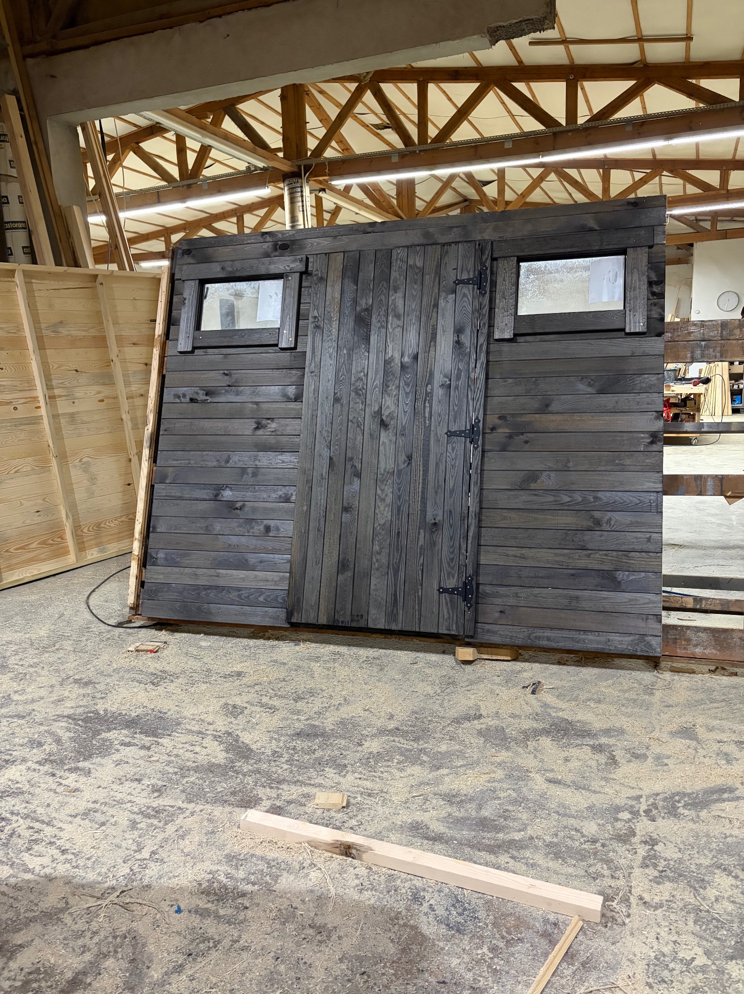 Ciemna, drewniana konstrukcja z drzwiami i oknami, prawdopodobnie fragment większej budowli, w warsztacie. Widoczne ślady obróbki drewna na podłodze.