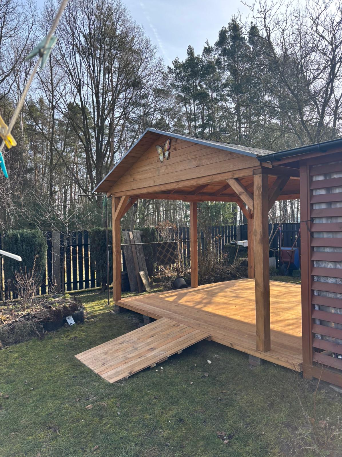 Drewniana altana z rampą wejściową, ozdobiona motylem na dachu, na tle ogrodu z drzewami i czarnym płotem. Słoneczny dzień.