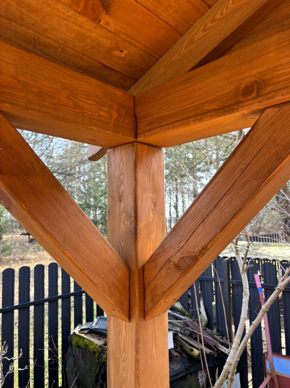 Detal drewnianej konstrukcji altany z widocznym słupem, belkami i fragmentem dachu. W tle zieleń i czarne ogrodzenie.