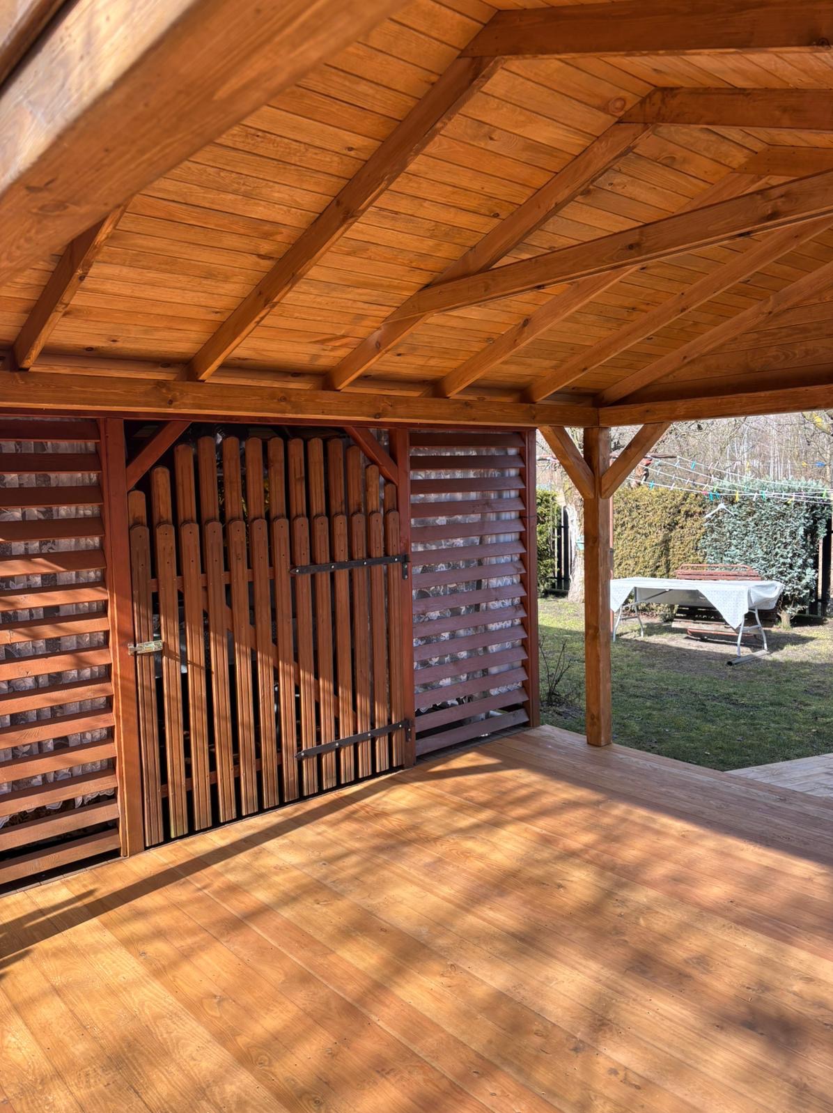 Zadaszona, drewniana konstrukcja z podłogą i ażurowymi ścianami, zamykana na drewniane wrota. W tle ogród z białym stołem i krzesłami.