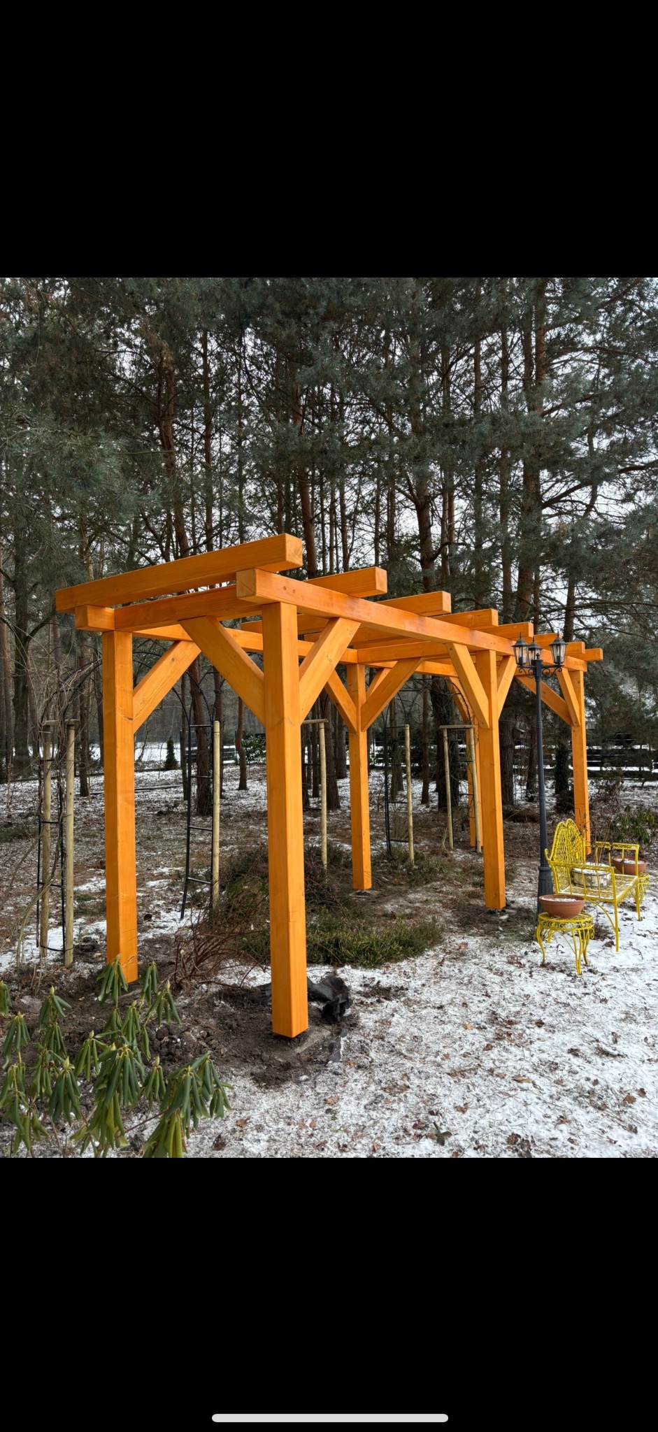 Nowa, pomarańczowa pergola w zimowym ogrodzie, z żółtą ławką i latarnią w tle, otoczona drzewami i śniegiem. Widok na konstrukcję w naturalnym otoczeniu.