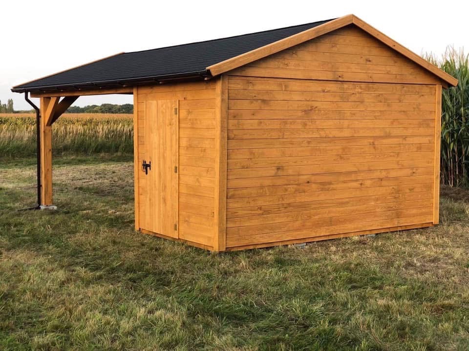 Drewniana altanka z czarnym dachem na trawie, z widocznym zadaszeniem wspartym na słupach. Naturalne oświetlenie, tło z polami i roślinnością.