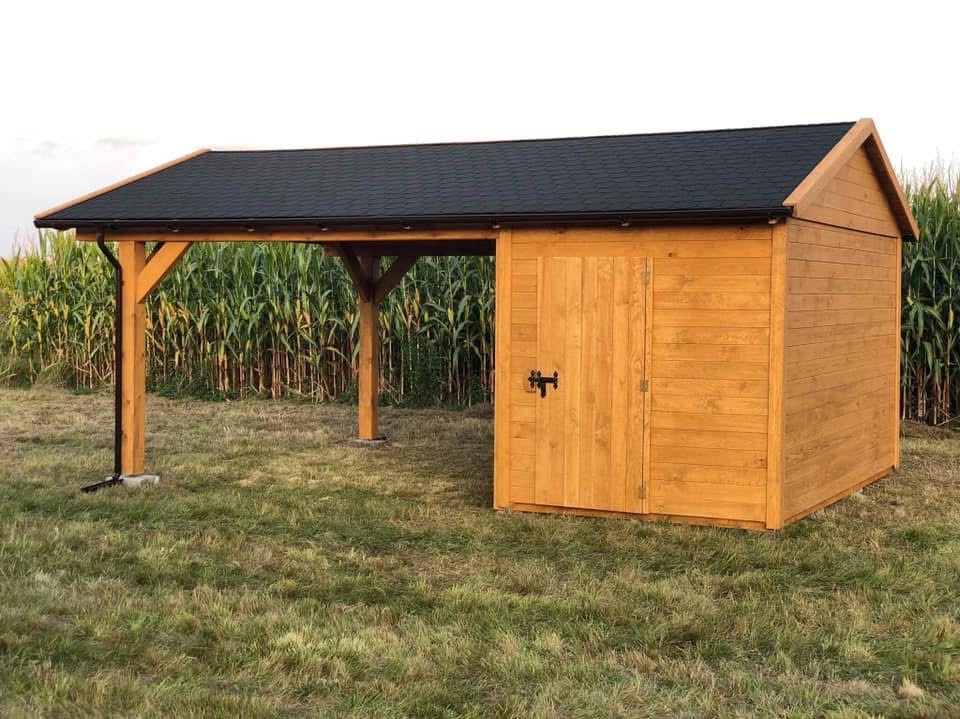 Drewniana konstrukcja z zadaszeniem i pomieszczeniem gospodarczym na trawie, z kukurydzą w tle. Całość w ciepłym odcieniu drewna, z czarnym dachem.