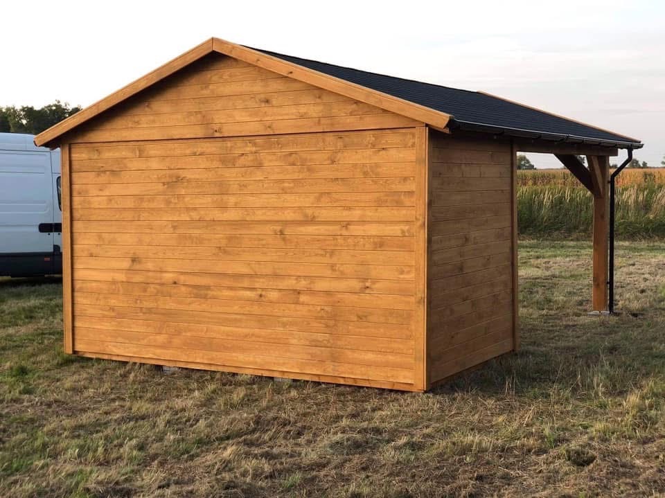 Drewniana konstrukcja z zadaszeniem na trawiastym terenie, widoczny fragment białego samochodu dostawczego w tle. Całość w ciepłej tonacji, równomierne oświetlenie.