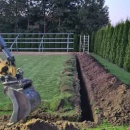 Wykopany wąski rów w trawie, obok sterta ziemi i ramię koparki. W tle rząd tuj i metalowa konstrukcja.